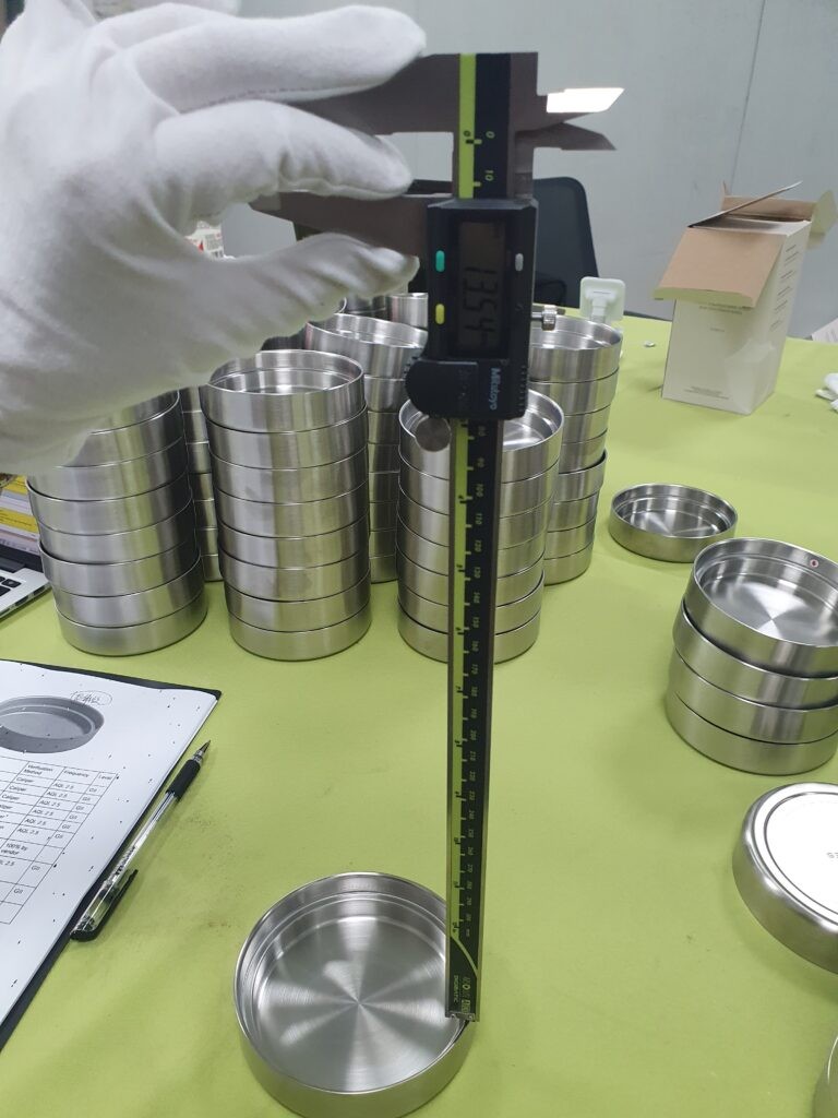 Gloved hand measuring a stainless steel lid with a digital caliper during an inspection